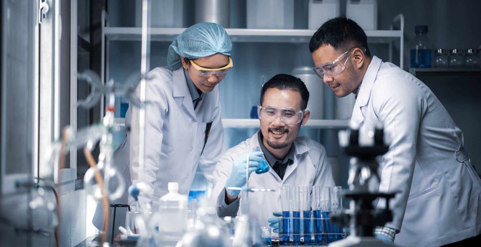 Three lab technicians wearing protective glasses and hair nets, working amid lab equipment