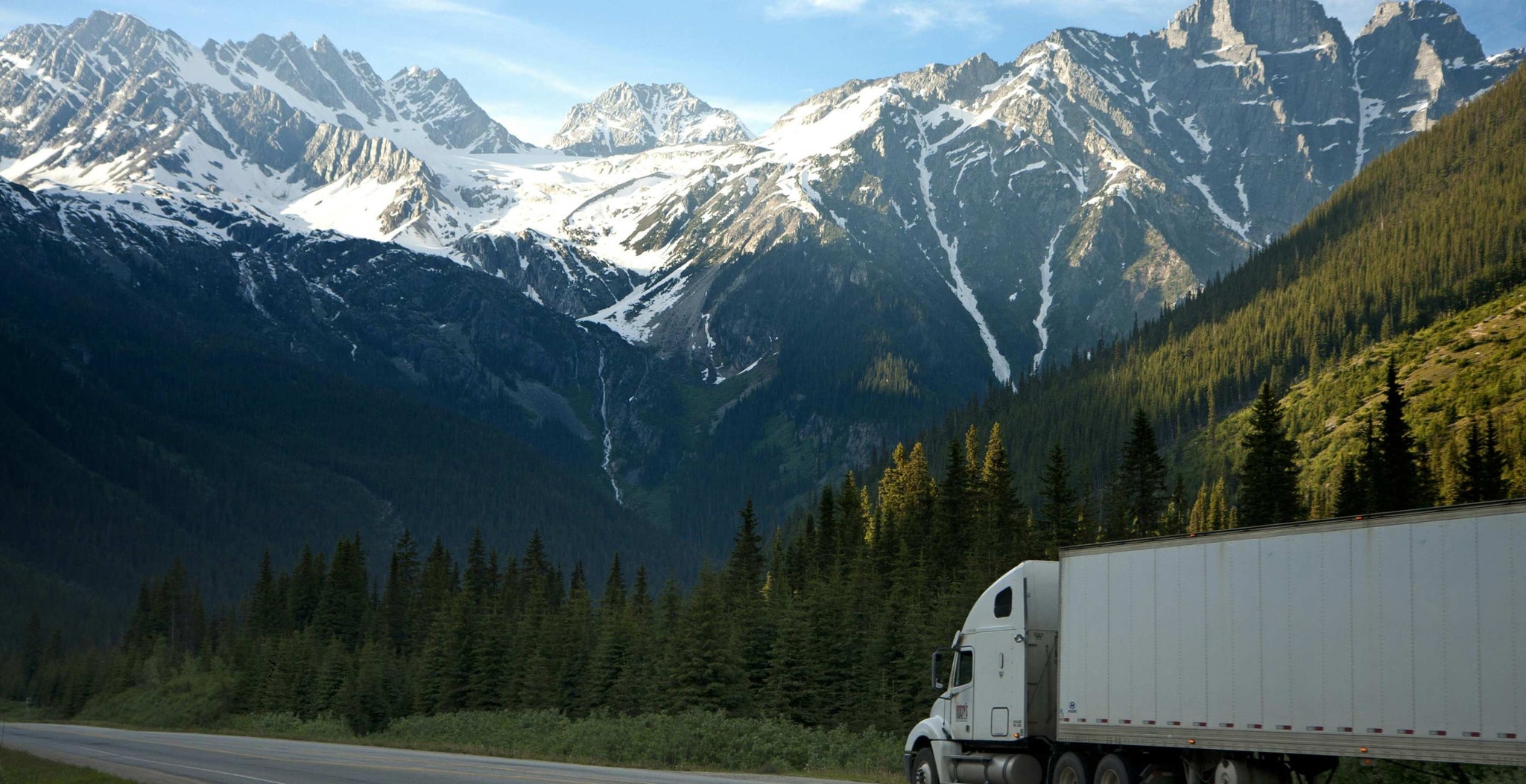 Truck on a mountain road with snow-capped peaks and green trees.