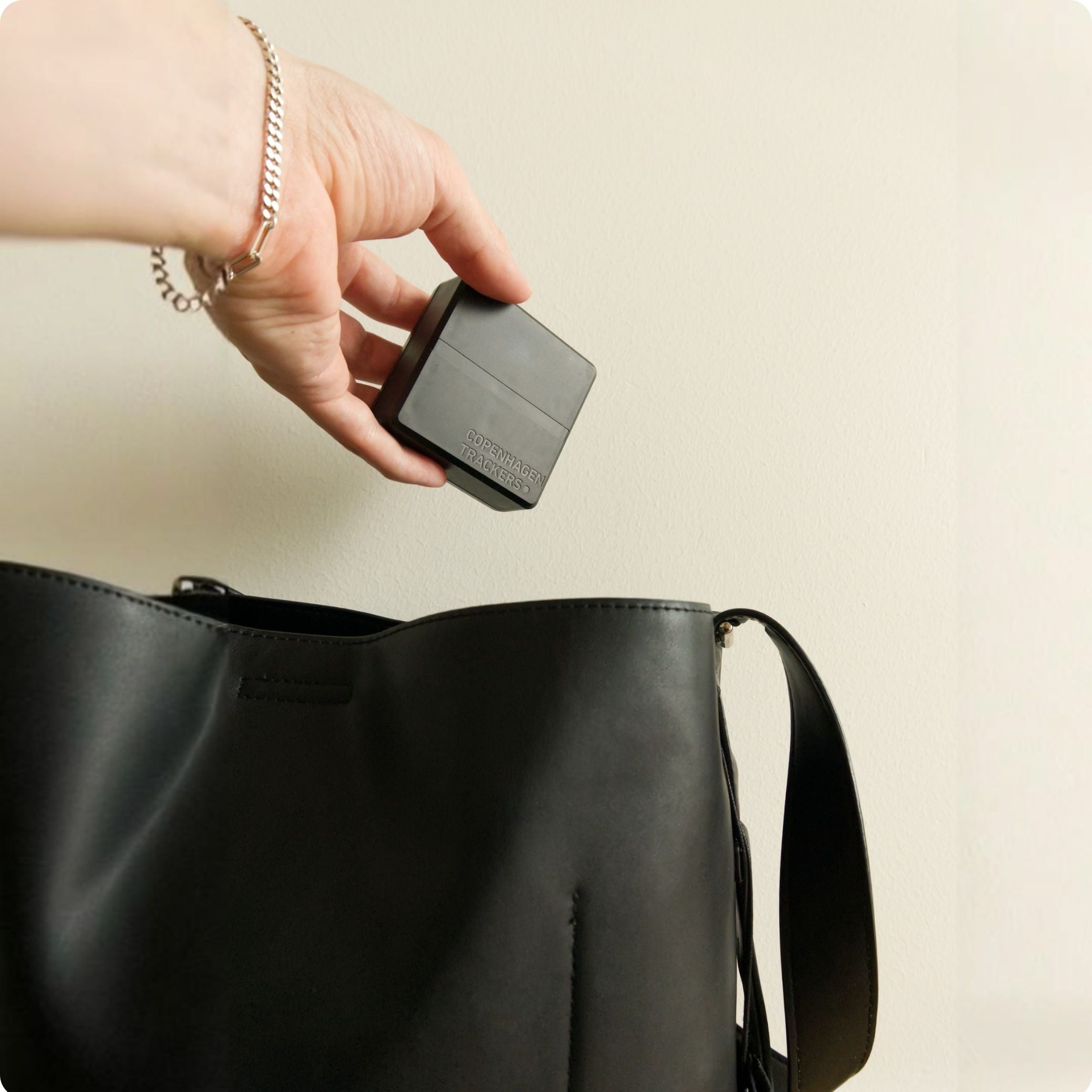 Mano che regge il localizzatore GPS Cobblestone 2.0 sopra una borsa nera su uno sfondo beige