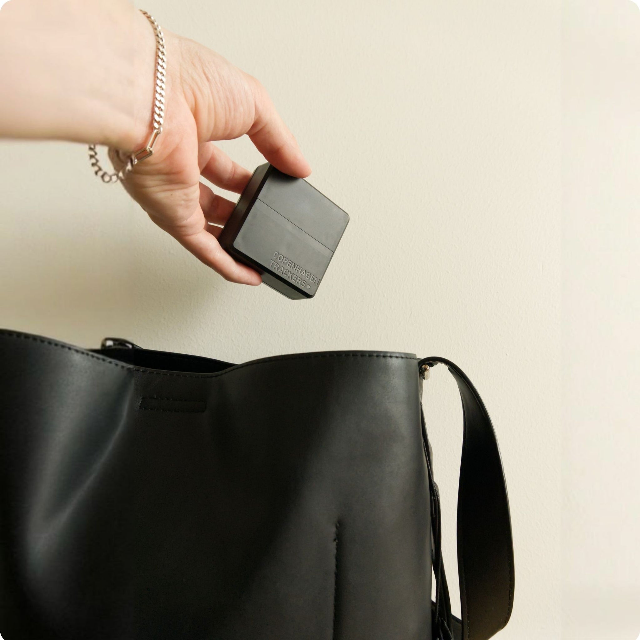 Mano che regge il localizzatore GPS Cobblestone 2.0 sopra una borsa nera su uno sfondo beige
