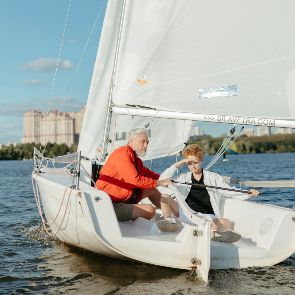Großvater und sein Enkel auf einem kleinen Segelboot an einem sonnigen Tag mit der Stadt im Hintergrund