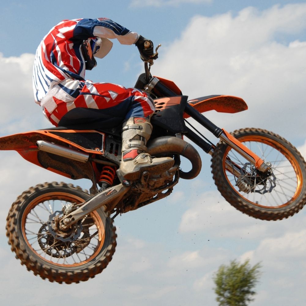 Persona en una moto de cross naranja realizando una acrobacia contra un cielo azul con nubes.