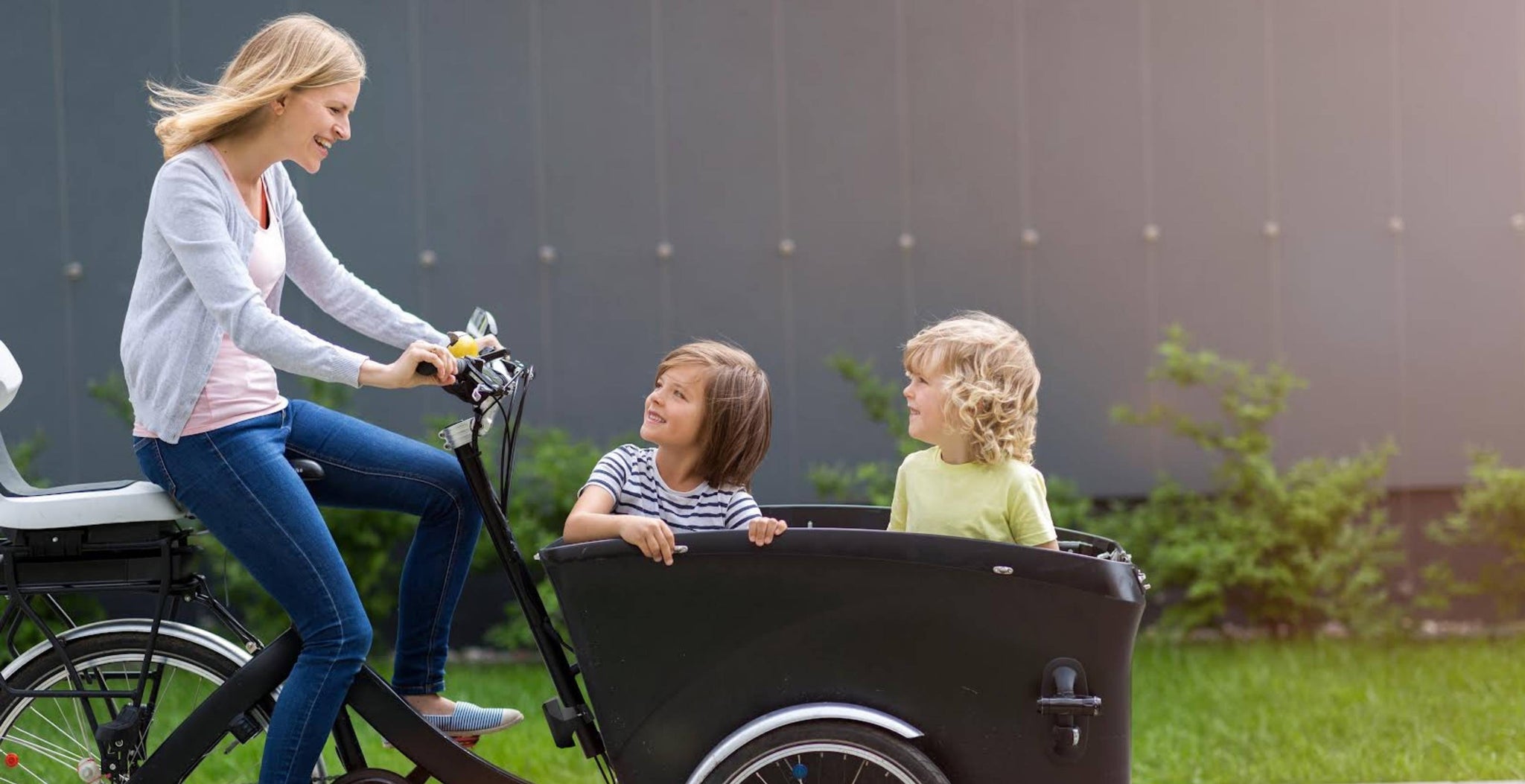 Mère souriante roulant sur un vélo cargo à travers la ville avec ses deux fils à l'avant qui la regardent en arrière, avec de la verdure en arrière-plan.