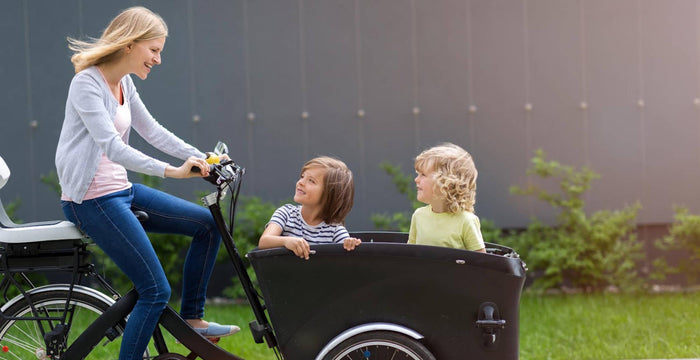 Mère souriante roulant sur un vélo cargo à travers la ville avec ses deux fils à l'avant qui la regardent en arrière, avec de la verdure en arrière-plan.